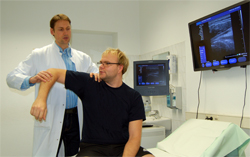 Arzt und Patient in schwarzem T-Shirt mit angewinkeltem Arm in der Orthopädie der Uniklinik Rostock