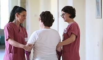 zwei junge Frauen in rosa Blusen führen Frau über den Gang in der Orthopädie Rostock